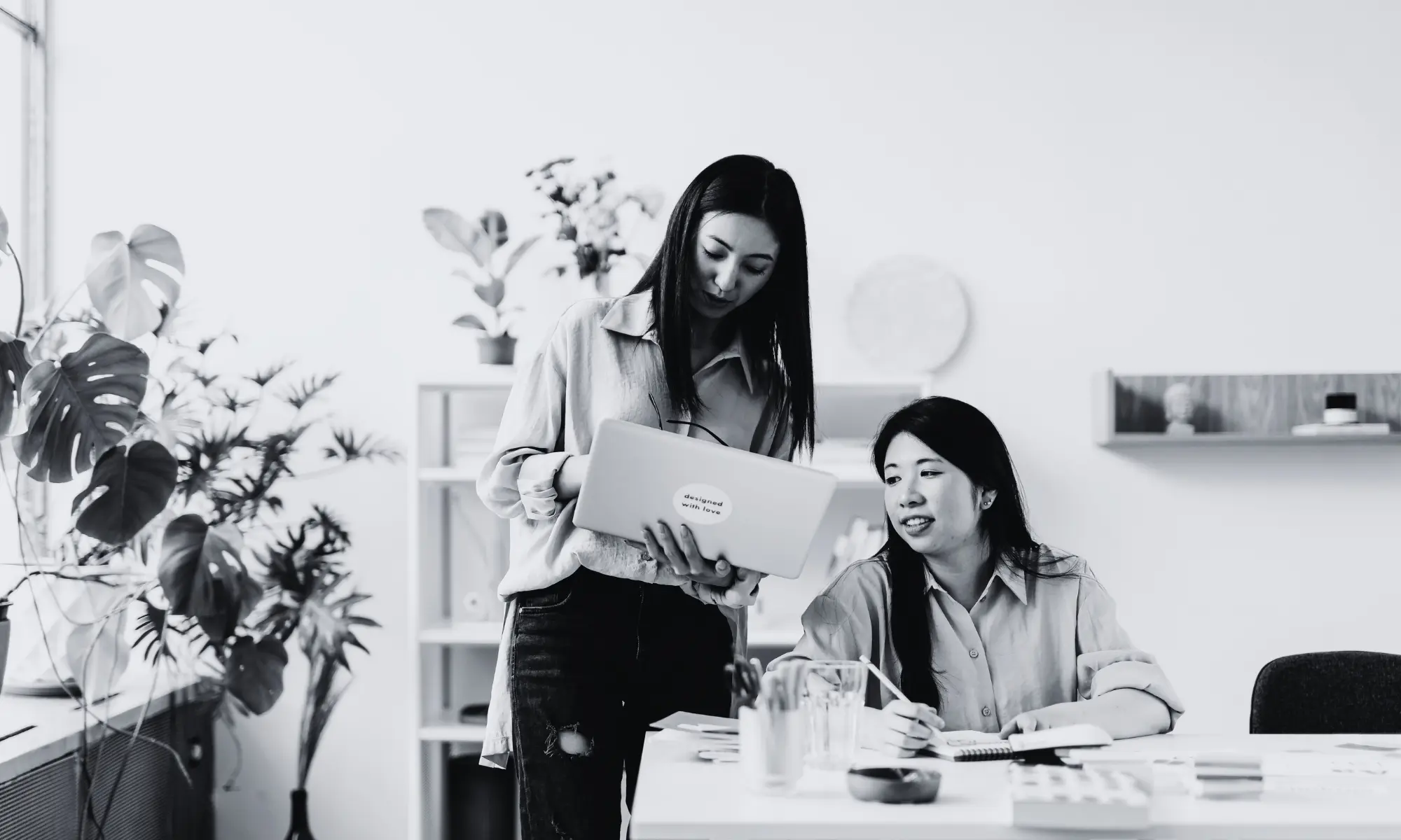 Two women discussing a laptop in a bright office. One stands holding the laptop; the other sits at a desk.